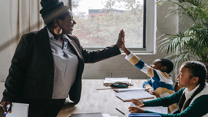 Teacher giving a high-five to a student in a classroom, with another student seated at a table with school supplies.